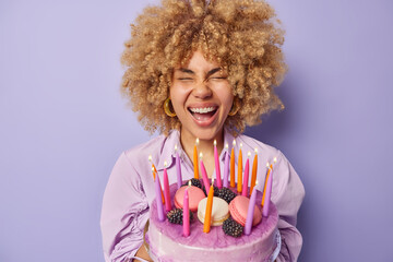 Canvas Print - Photo of curly haired young woman poses with delicious cake decorated with macaroons exclaims loudly enjoys celebration wears earrings and shirt isolated over purple background. Holiday concept