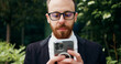 © serg - Close up. Cheerful man in glasses sitting on bench outdoors using typing smartphone. Confident bearded male resting on bench near office building texting at mobile phone.