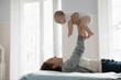 Side view loving mother lying on comfortable bed in cozy, light bedroom play with infant, lifts un outstretched arms her adorable awakened newborn smile, enjoy sweet moments together at home. Infancy