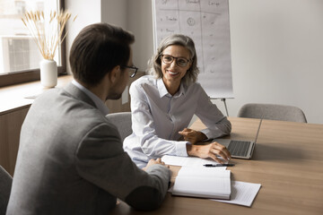 Sticker - Happy mature businesswoman talking to younger male colleague at laptop, smiling, laughing, discussing work project cooperation in meeting room, brainstorming on ideas