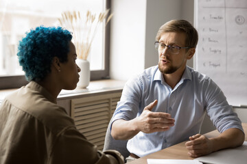 Wall Mural - Two multiethnic coworkers talking at office meeting table, discussing work process, collaboration, partnership, planning teamwork. Young business leader man, boss explaining task to colleague woman