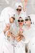 © wedmoments.stock - Young, beautiful women in bathrobes and glasses enjoying bachelorette party at spa or hotel and eating donuts, happy multiracial girlfriends celebrating bachelorette party