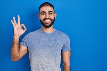 Wall Mural - Middle east man with beard standing over blue background smiling positive doing ok sign with hand and fingers. successful expression.