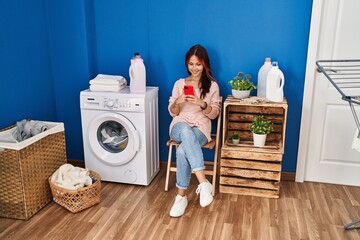 Wall Mural - Young caucasian woman using smartphone waiting for washing machine at laundry room