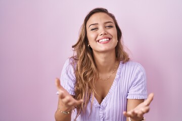 Wall Mural - Young hispanic woman standing over pink background smiling cheerful offering hands giving assistance and acceptance.