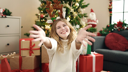 Sticker - Young blonde woman sitting on floor by christmas tree with arms open at home