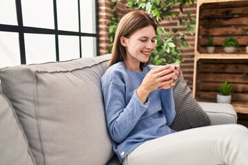 Wall Mural - Young blonde woman drinking coffee sitting on sofa at home