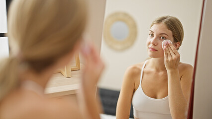 Wall Mural - Young blonde woman cleaning face with cotton pad looking on mirror at home