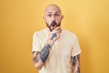 Poster - Hispanic man with tattoos standing over yellow background looking fascinated with disbelief, surprise and amazed expression with hands on chin