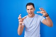 © Krakenimages.com - Young hispanic man standing over blue background shouting frustrated with rage, hands trying to strangle, yelling mad