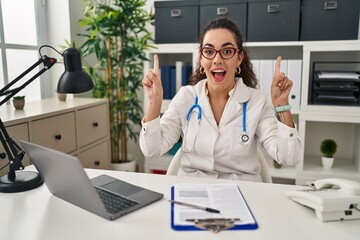 Canvas Print - Young hispanic woman wearing doctor uniform and stethoscope smiling amazed and surprised and pointing up with fingers and raised arms.