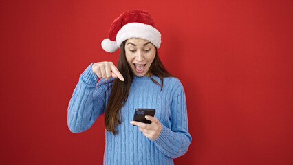 Sticker - Young caucasian woman wearing christmas hat pointing to smartphone over isolated red background