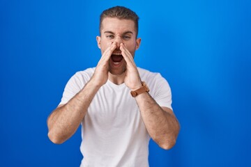 Poster - Young caucasian man standing over blue background shouting angry out loud with hands over mouth