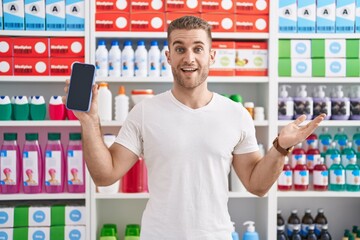 Wall Mural - Young caucasian man working at pharmacy drugstore showing smartphone screen celebrating achievement with happy smile and winner expression with raised hand