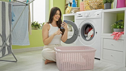 Wall Mural - Young beautiful hispanic woman washing clothes using smartphone at laundry room