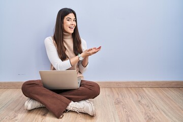 Canvas Print - Young brunette woman working using computer laptop sitting on the floor pointing aside with hands open palms showing copy space, presenting advertisement smiling excited happy