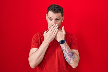 Poster - Young hispanic man standing over red background shocked covering mouth with hands for mistake. secret concept.