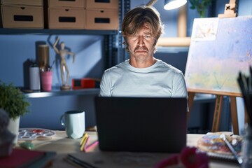 Wall Mural - Middle age man sitting at art studio with laptop at night depressed and worry for distress, crying angry and afraid. sad expression.