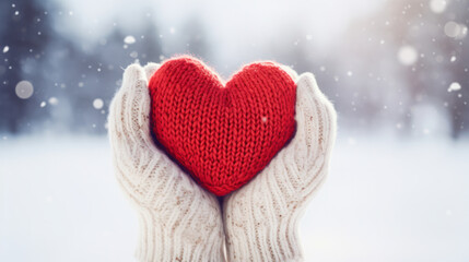 Close-up of female hands in mittens holding a beautiful knitted heart on a snowy winter background. Concept of love, relaxation, cozy.