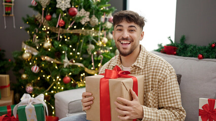 Wall Mural - Young arab man holding gift sitting by christmas tree at home