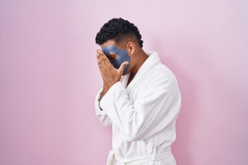 Sticker - Young hispanic man wearing beauty face mask and bath robe with sad expression covering face with hands while crying. depression concept.