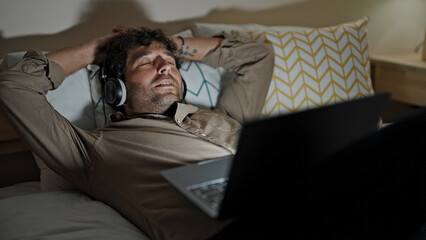 Canvas Print - Young hispanic man using laptop and headphones relaxed on bed at bedroom