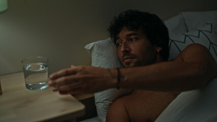 Wall Mural - Young hispanic man holding glass of water lying on bed shirtless at bedroom