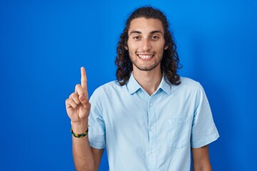 Sticker - Young hispanic man standing over blue background showing and pointing up with finger number one while smiling confident and happy.