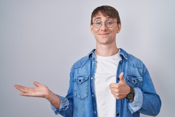 Wall Mural - Caucasian blond man standing wearing glasses showing palm hand and doing ok gesture with thumbs up, smiling happy and cheerful