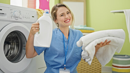 Canvas Print - Young blonde woman professional cleaner holding detergent bottle and folded towels at laundry room