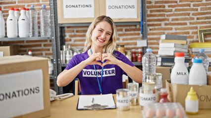 Poster - Young blonde woman volunteer sitting on table doing heart gesture at charity center