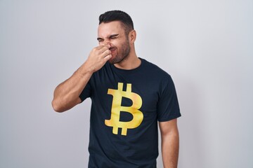 Poster - Young hispanic man wearing bitcoin t shirt smelling something stinky and disgusting, intolerable smell, holding breath with fingers on nose. bad smell