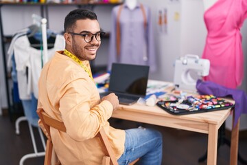 Wall Mural - Young hispanic man tailor smiling confident using laptop at tailor shop