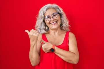 Canvas Print - Middle age woman with grey hair standing over red background pointing to the back behind with hand and thumbs up, smiling confident