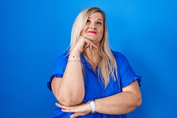 Wall Mural - Caucasian plus size woman standing over blue background with hand on chin thinking about question, pensive expression. smiling with thoughtful face. doubt concept.