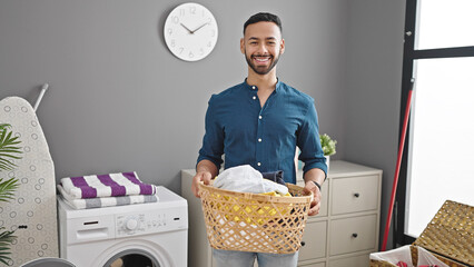 Wall Mural - Young hispanic man smiling confident holding basket with clothes at laundry room