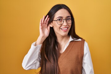 Poster - Young brunette woman standing over yellow background wearing glasses smiling with hand over ear listening an hearing to rumor or gossip. deafness concept.