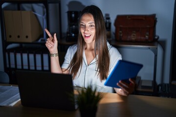 Canvas Print - Young brunette woman working at the office at night with a big smile on face, pointing with hand finger to the side looking at the camera.