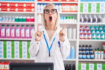 Wall Mural - Young caucasian woman working at pharmacy drugstore amazed and surprised looking up and pointing with fingers and raised arms.