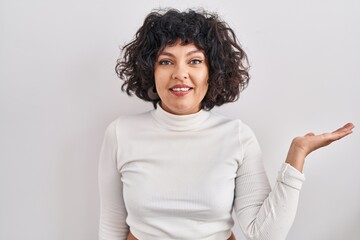 Poster - Hispanic woman with curly hair standing over isolated background smiling cheerful presenting and pointing with palm of hand looking at the camera.