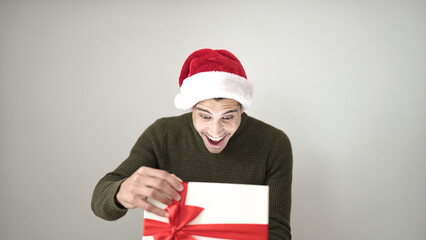 Sticker - Young hispanic man unpacking gift wearing christmas hat over isolated white background