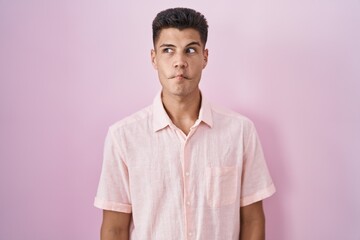 Wall Mural - Young hispanic man standing over pink background making fish face with lips, crazy and comical gesture. funny expression.