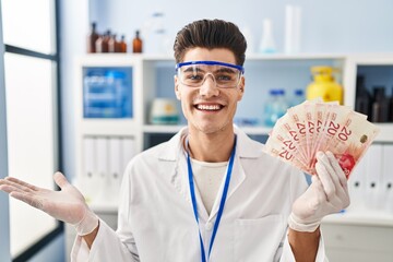 Wall Mural - Young hispanic man working at scientist laboratory holding shekels celebrating achievement with happy smile and winner expression with raised hand