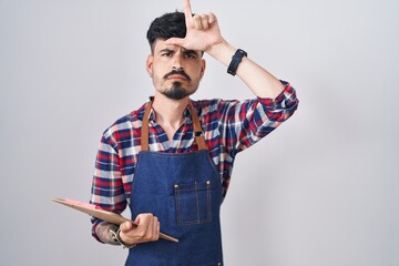 Sticker - Young hispanic man with beard wearing waiter apron holding clipboard making fun of people with fingers on forehead doing loser gesture mocking and insulting.