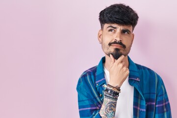 Wall Mural - Young hispanic man with beard standing over pink background looking confident at the camera with smile with crossed arms and hand raised on chin. thinking positive.