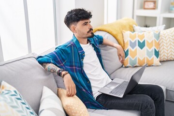 Wall Mural - Young hispanic man using laptop sitting on sofa at home