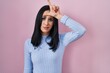 Hispanic woman standing over pink background making fun of people with fingers on forehead doing loser gesture mocking and insulting.