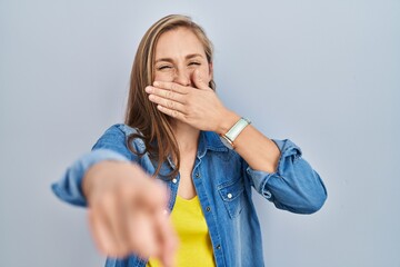 Sticker - Young blonde woman standing over blue background laughing at you, pointing finger to the camera with hand over mouth, shame expression