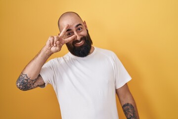 Canvas Print - Young hispanic man with beard and tattoos standing over yellow background doing peace symbol with fingers over face, smiling cheerful showing victory