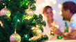 Árvore de Natal em primeiro plano com duas pessoas sorridentes desfocadas ao fundo.